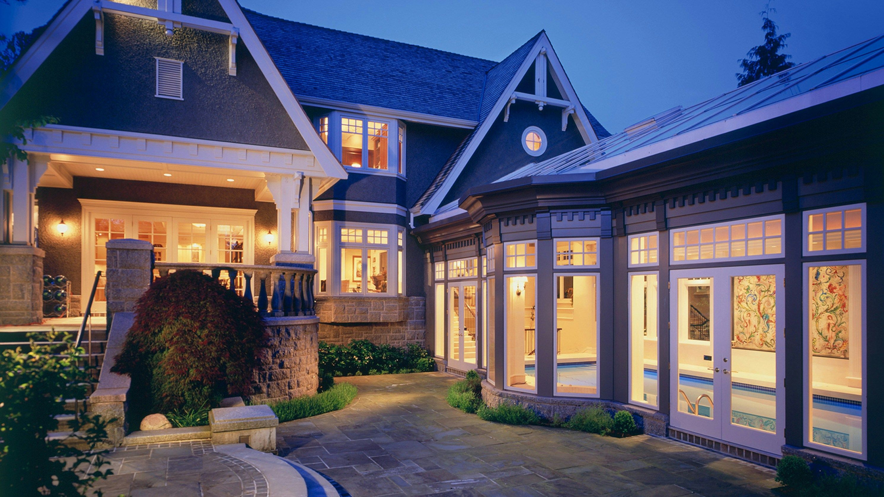 A two-story suburban house at dusk with smart lighting simulation: a warm porch light is on, upstairs bedroom lights are glowing, and a flickering blue light in the ground floor window mimics the glow of a television to create a lived-in look.