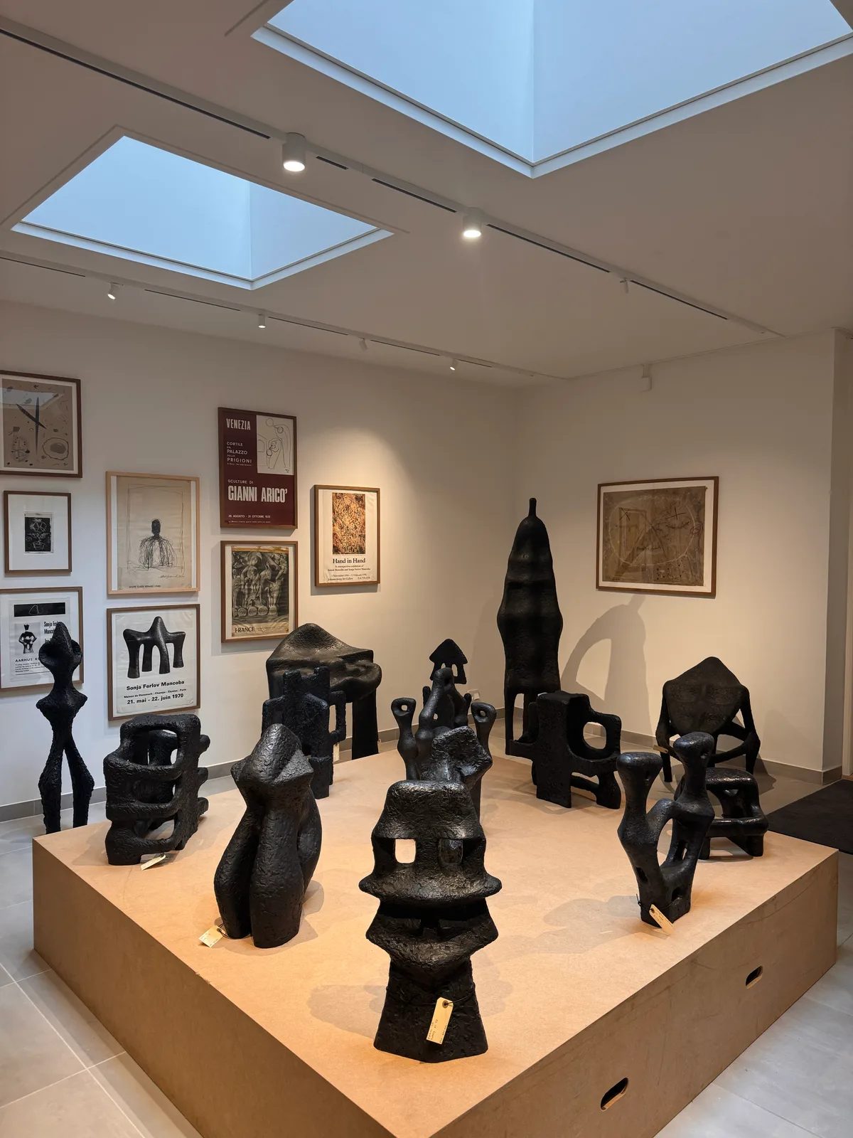 Dark textured sculptures on a wooden display platform beneath skylights, with framed exhibition posters and drawings on the walls.