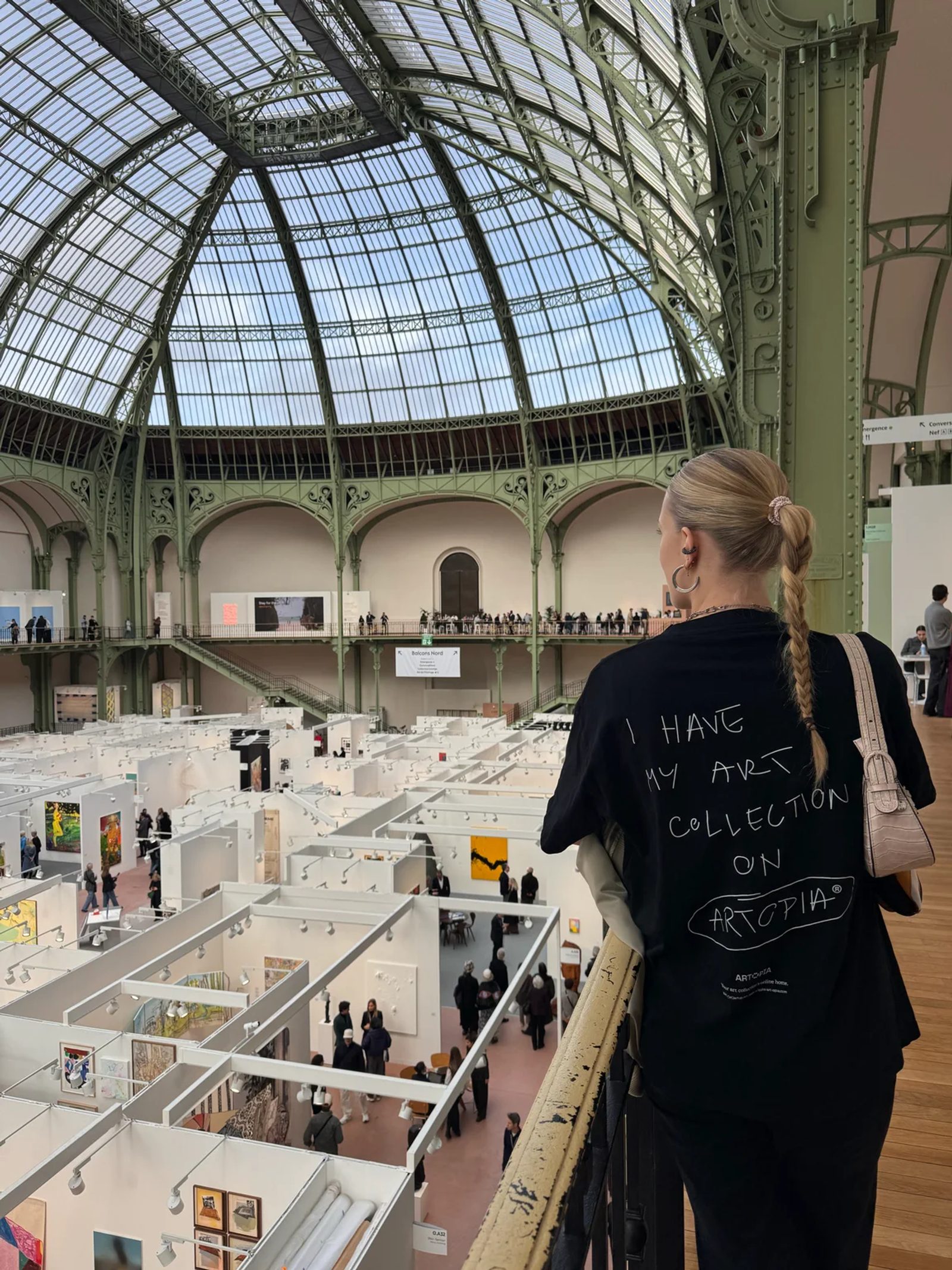A visitor wearing an Artopia T-shirt looks over Art Basel Paris booths from a balcony beneath the Grand Palais glass roof.