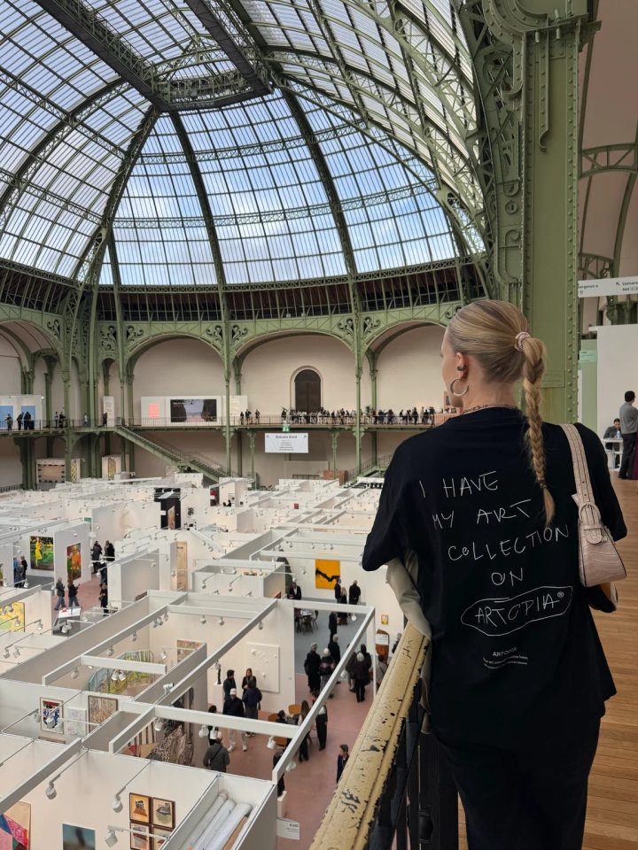 A visitor wearing an Artopia T-shirt looks over Art Basel Paris booths from a balcony beneath the Grand Palais glass roof.