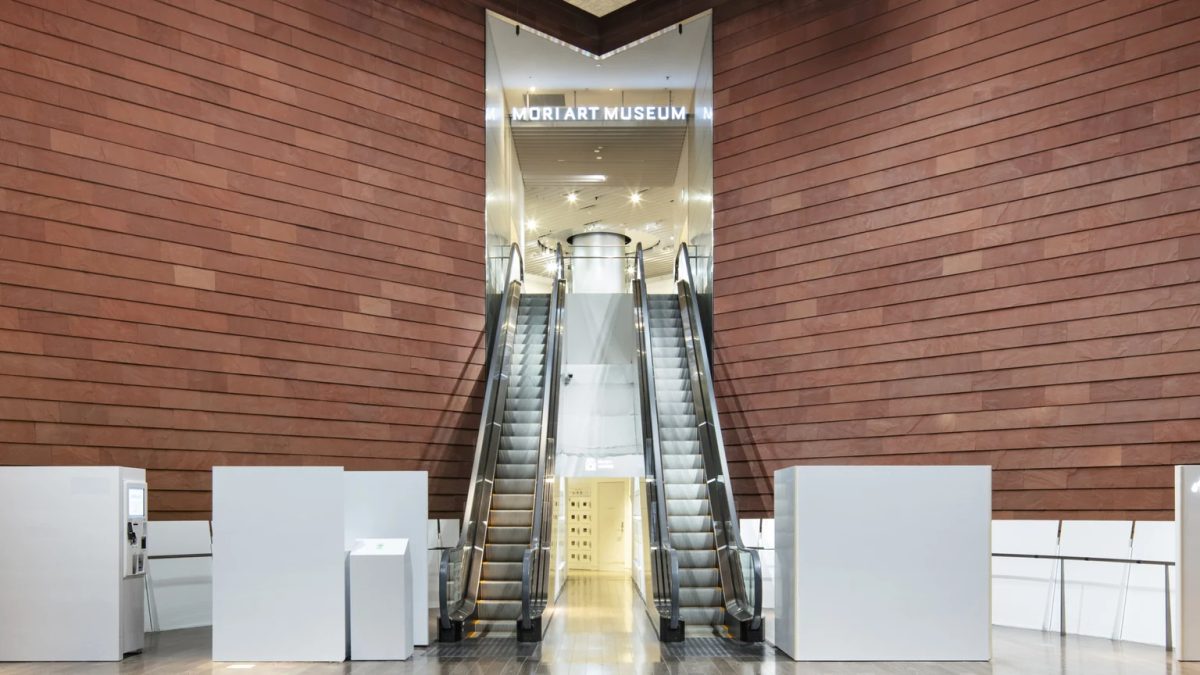 Escalators leading into the Mori Art Museum between warm red walls beneath the museum’s illuminated sign.