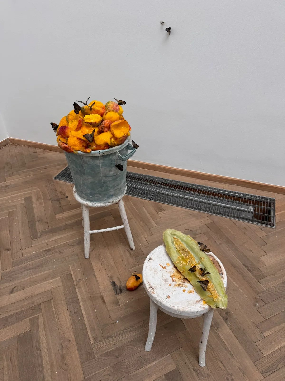 Yellow and orange fruit sculptures in a metal bucket beside a halved green melon sculpture on white stools, with butterflies.