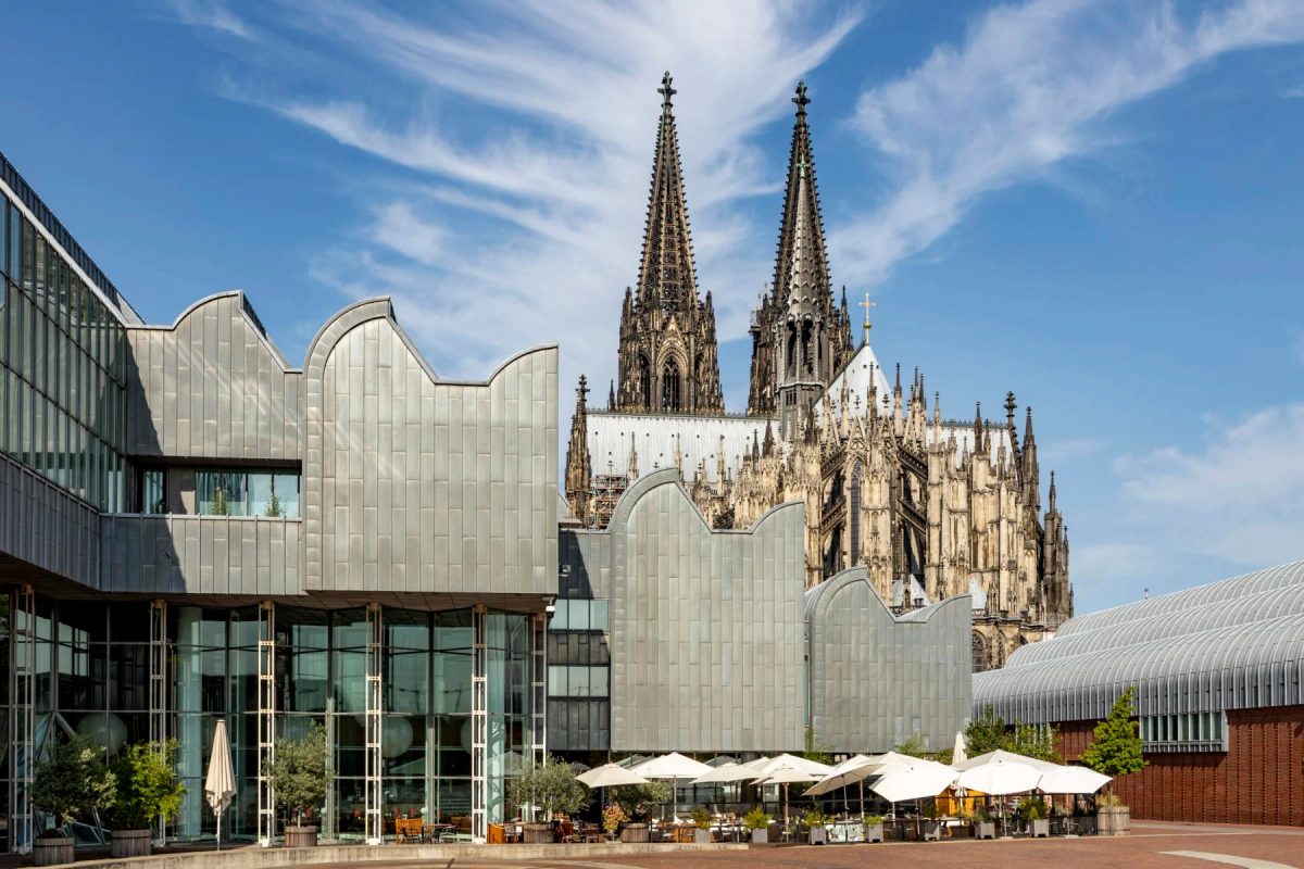 Museum Ludwig’s curved metal roofs and glass facade, with Cologne Cathedral rising behind the museum under a blue sky.