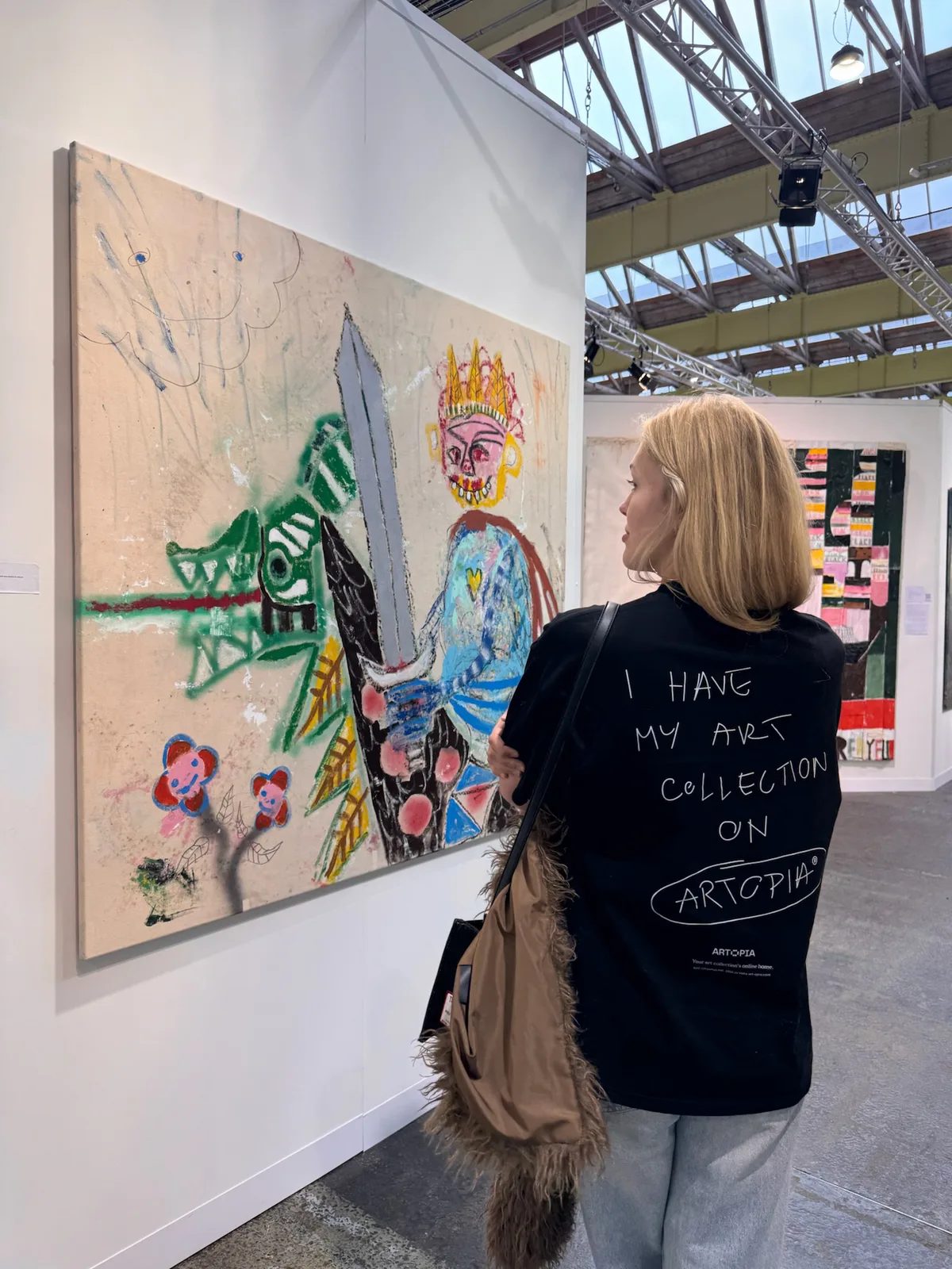 A visitor wearing an Artopia T-shirt stands beside a colourful painting by Niall Campbell Strachan at Enter Art Fair.