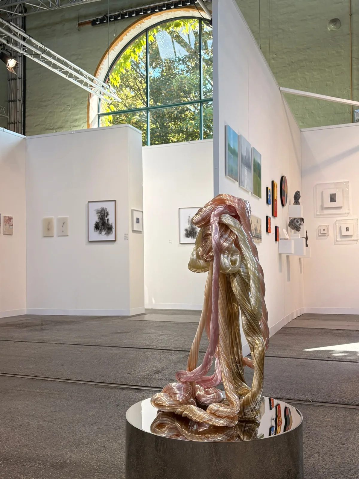A flowing pink and gold glass sculpture on a mirrored plinth in a bright art fair booth.