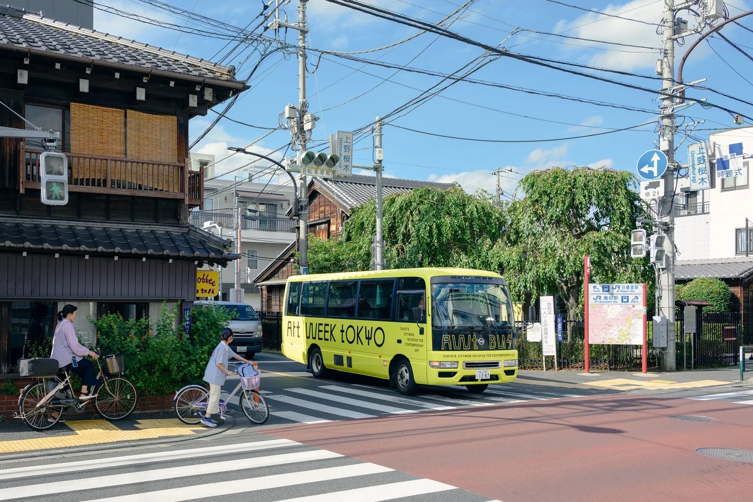 A lime-green Art Week Tokyo bus passing traditional wooden buildings and cyclists on a Tokyo street.