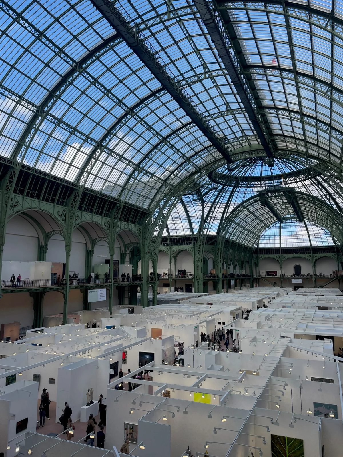 Art fair booths and visitors beneath the glass and steel roof of the Grand Palais in Paris.