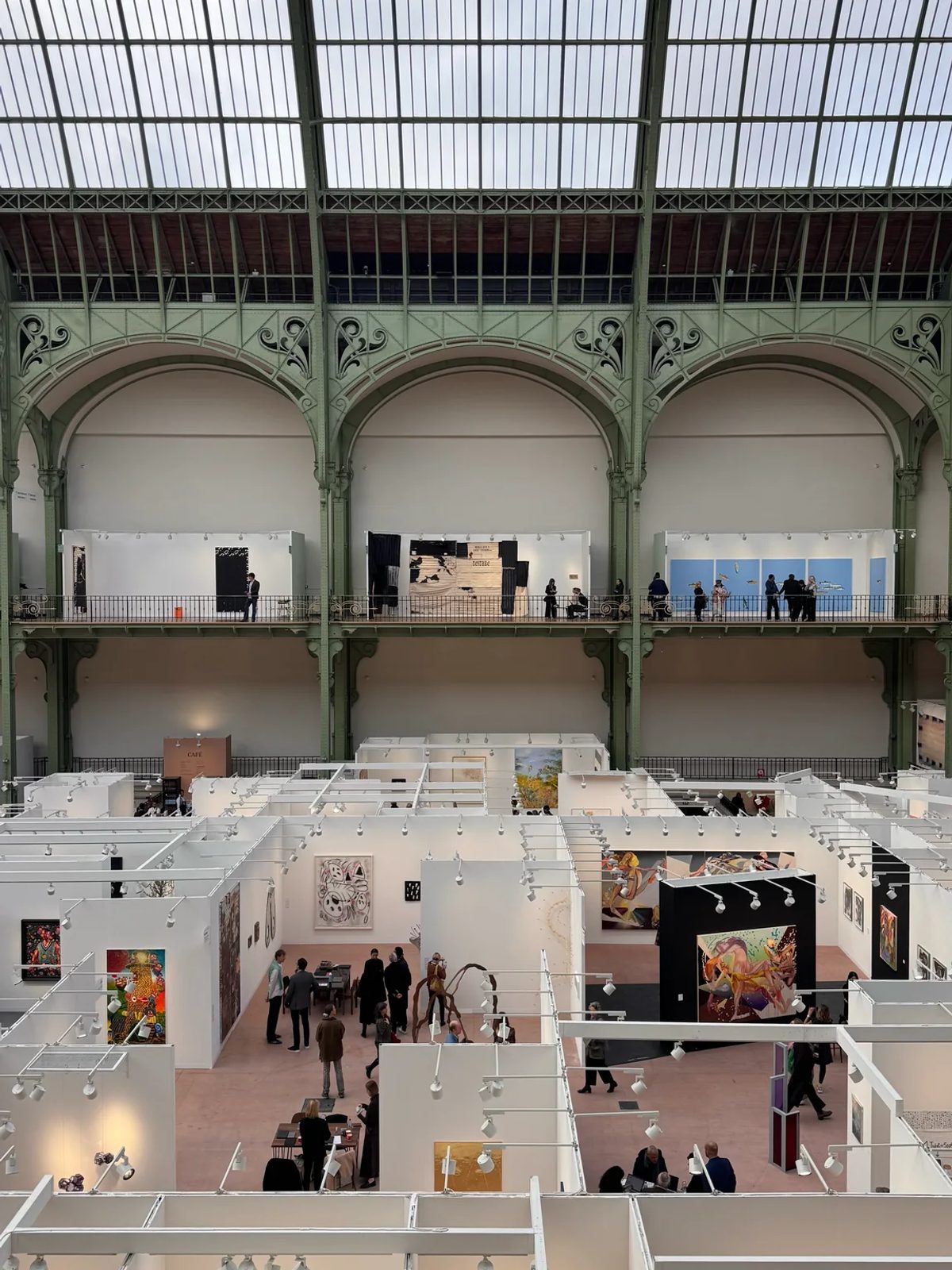 Art fair booths and visitors beneath the glass roof and green iron arches of the Grand Palais at Art Basel Paris.