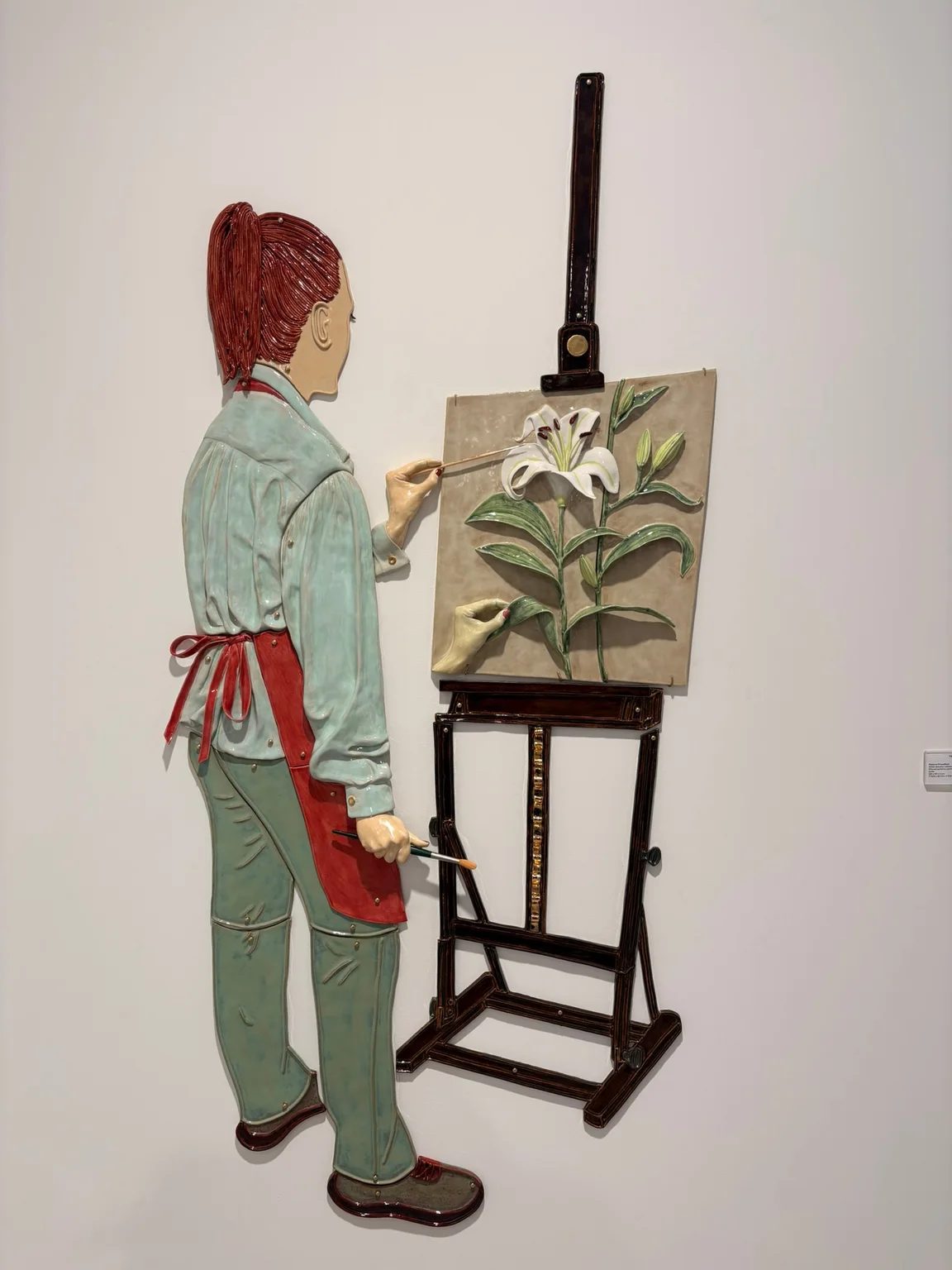 A ceramic wall relief by Paloma Proudfoot depicts a woman in a pale green suit painting a lily at an easel.