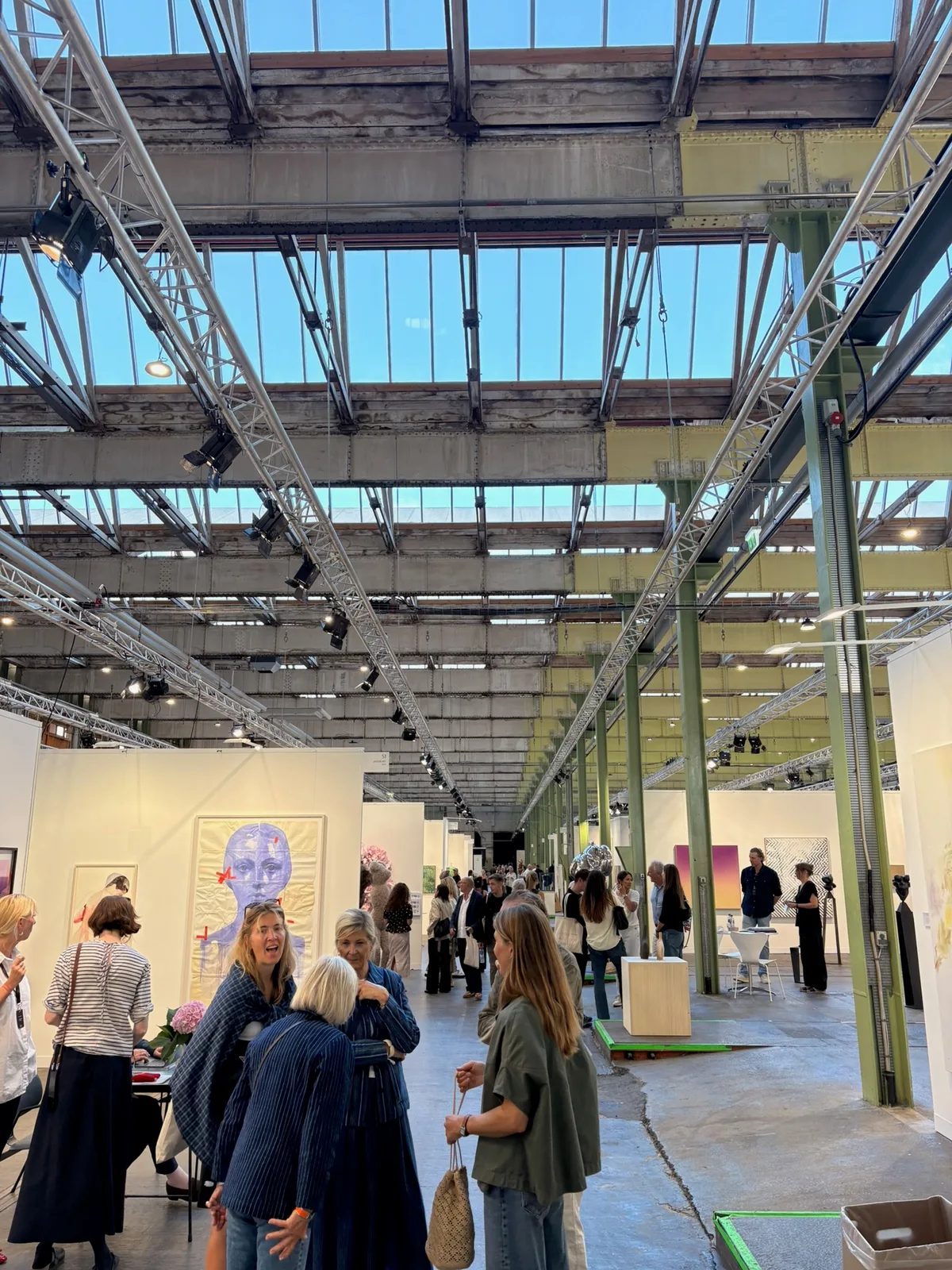 Visitors walking between art fair booths beneath the glass and steel roof of a former locomotive hall.