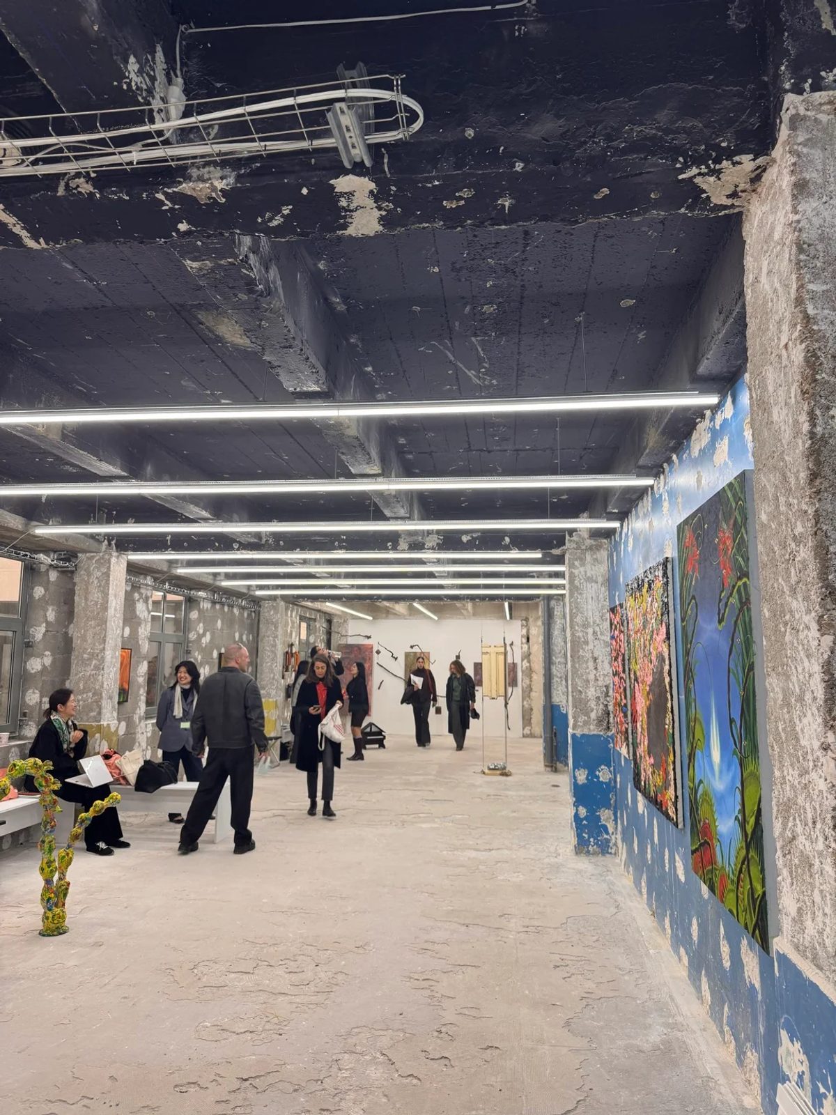 Visitors exploring paintings and sculptures inside Paris Internationale 2026, beside blue walls and exposed concrete columns.