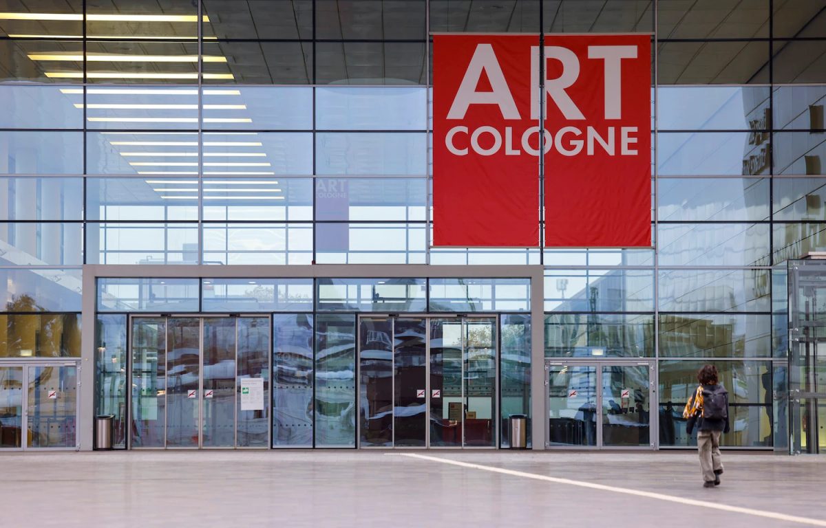 Entrance to Art Cologne at Koelnmesse, with a large red fair banner above the glass doors.