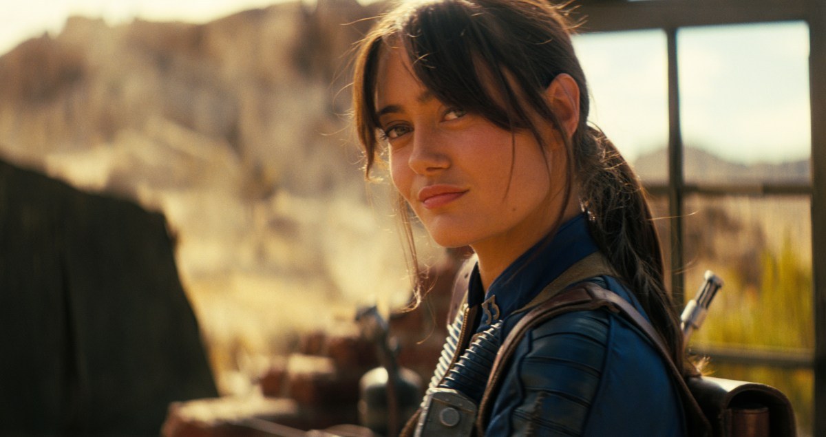 Lucy from the Fallout TV series looks back over her shoulder in a desert scene.