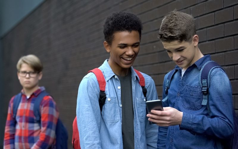 To gutter ser på en telefon og ler. En annen gutt står i bakgrunnen og føler seg utenfor
