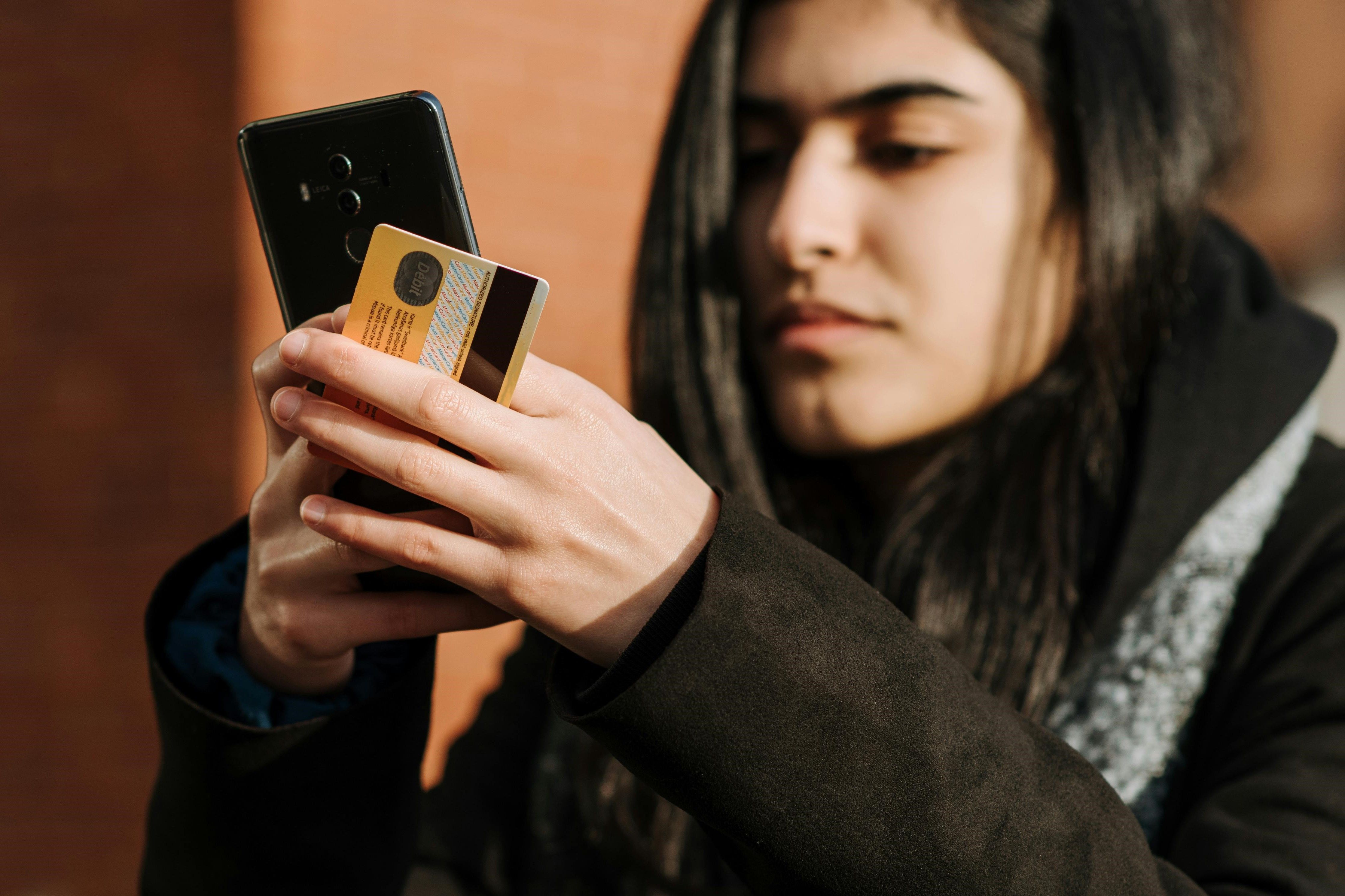 Tenåringsjente ordner noe på mobilen, mens hun holder et bankkort i den andre hånden.