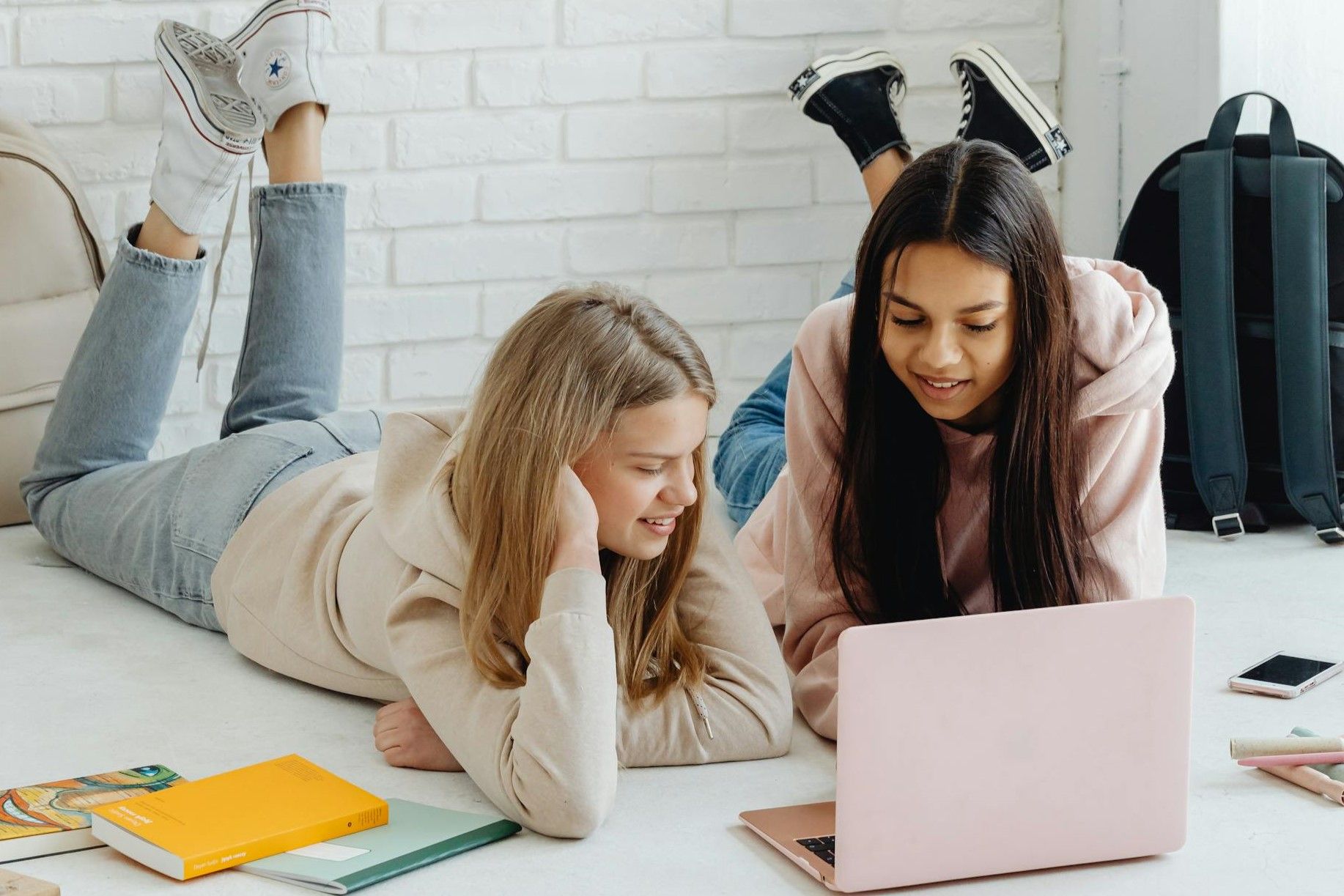 To tenåringsjenter ligger på magen på gulvet med en PC foran seg med skolebøker rundt.