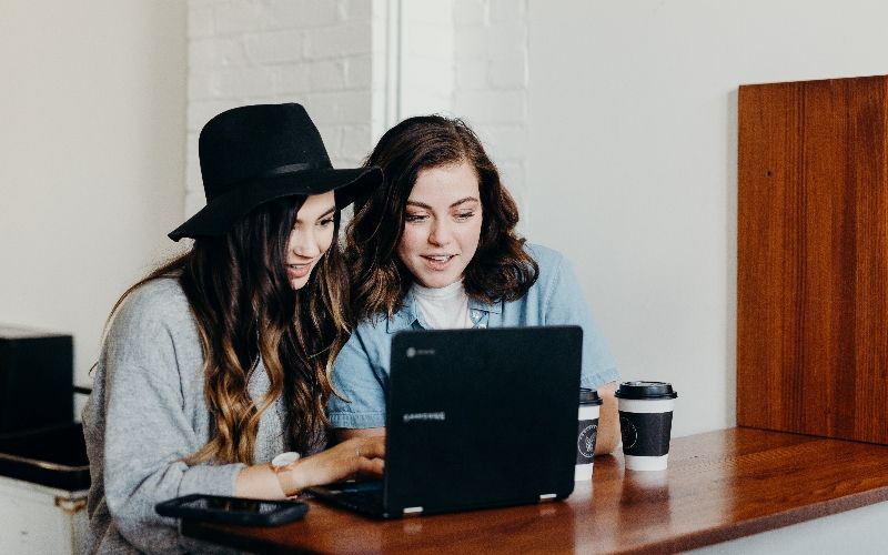 To smilende jenter bruker en datamaskin sammen. Foto: Unsplash / Brooke Cagle