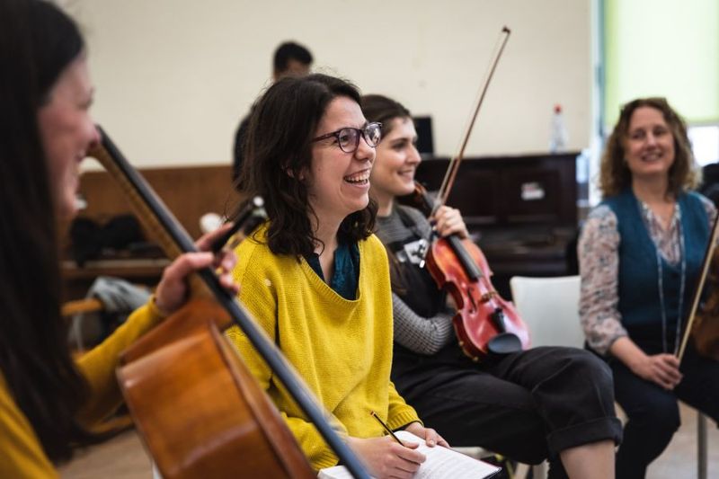 A Musicians of Change and music teacher laughing while holding a pen, surrounded by a strings ensemble in a school music room.