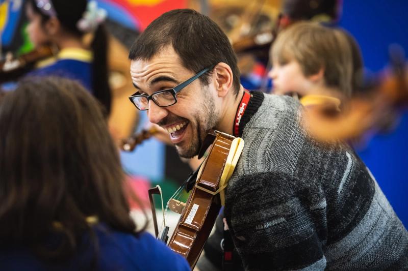 A Music Masters teacher smiling with his violin while leading a group of young string players.