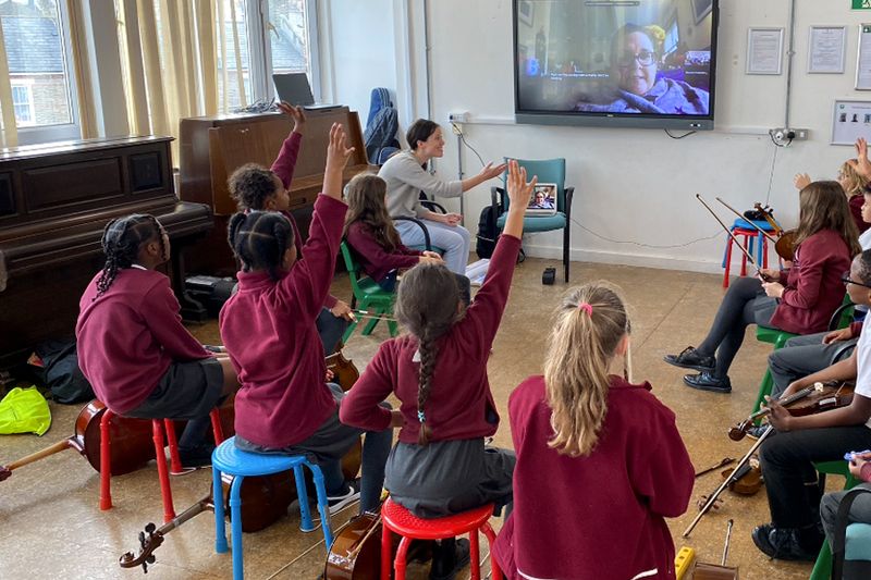Primary school children with their hands up, taking part in a workshop with a musician on a screen.