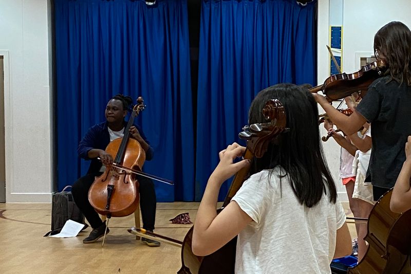 A cellist playing on a school hall stage while pupils watch and play along in the foreground.