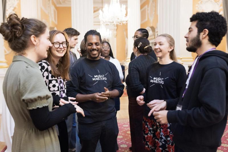 Music Masters staff and supporters in branded T-shirts talking and laughing together at an event in a grand room.