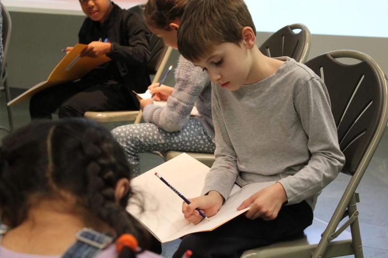 Children sitting on chairs in a workshop space, writing and drawing on paper resting on their laps.
