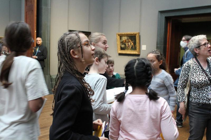 A group of children in a gallery looking up at a painting, with adults standing among them.