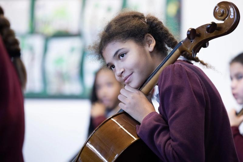 A girl in school uniform resting her head against the neck of her cello in a classroom.
