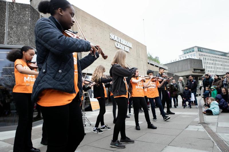 Young violinists in orange #SaveOurMusic T-shirts performing outdoors to a crowd at the Southbank Centre.