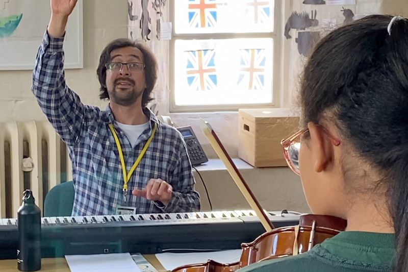A music leader with his arm raised conducting from behind a keyboard, a pupil with a cello in the foreground.
