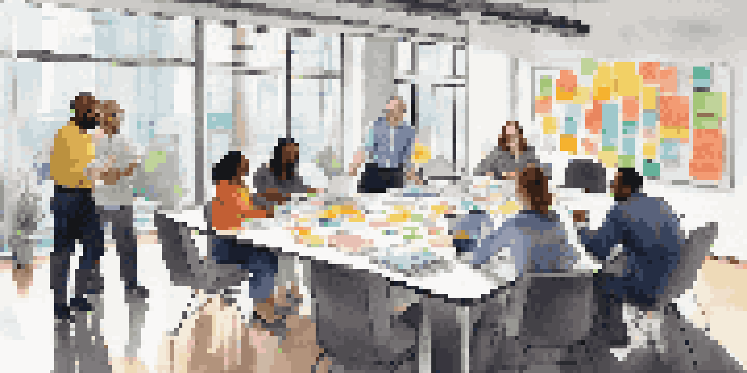 A diverse group of professionals collaborating at a table in a bright office, surrounded by laptops and sticky notes.
