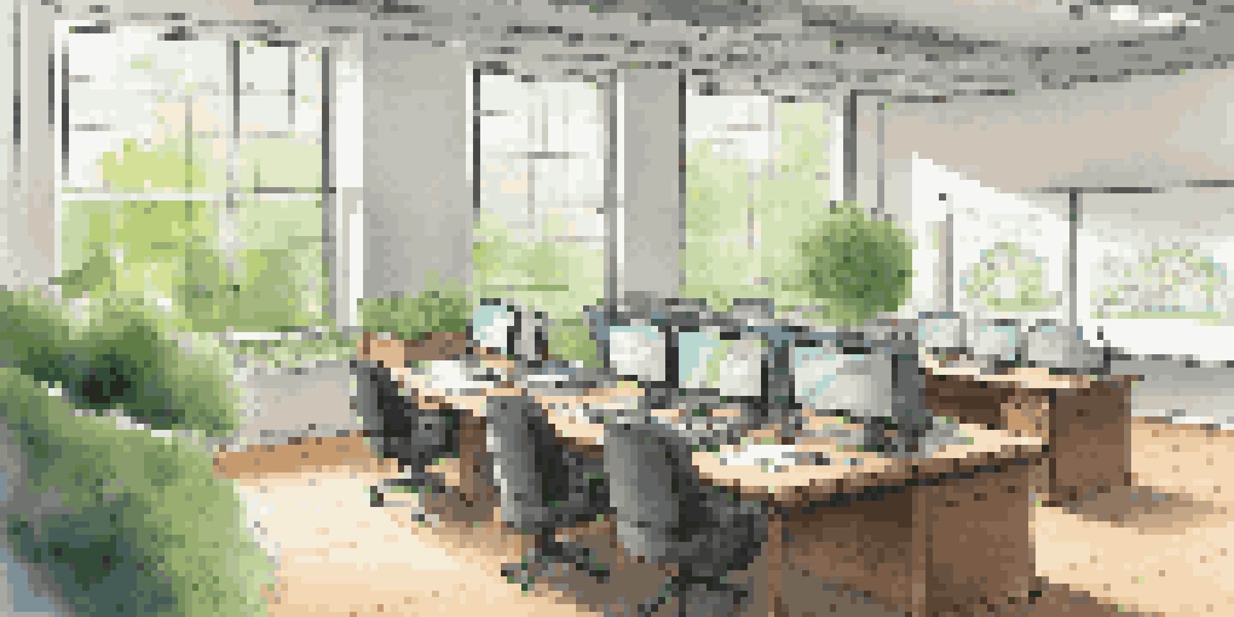 A bright and modern office space with a large table, laptops, and a whiteboard surrounded by greenery outside the windows.