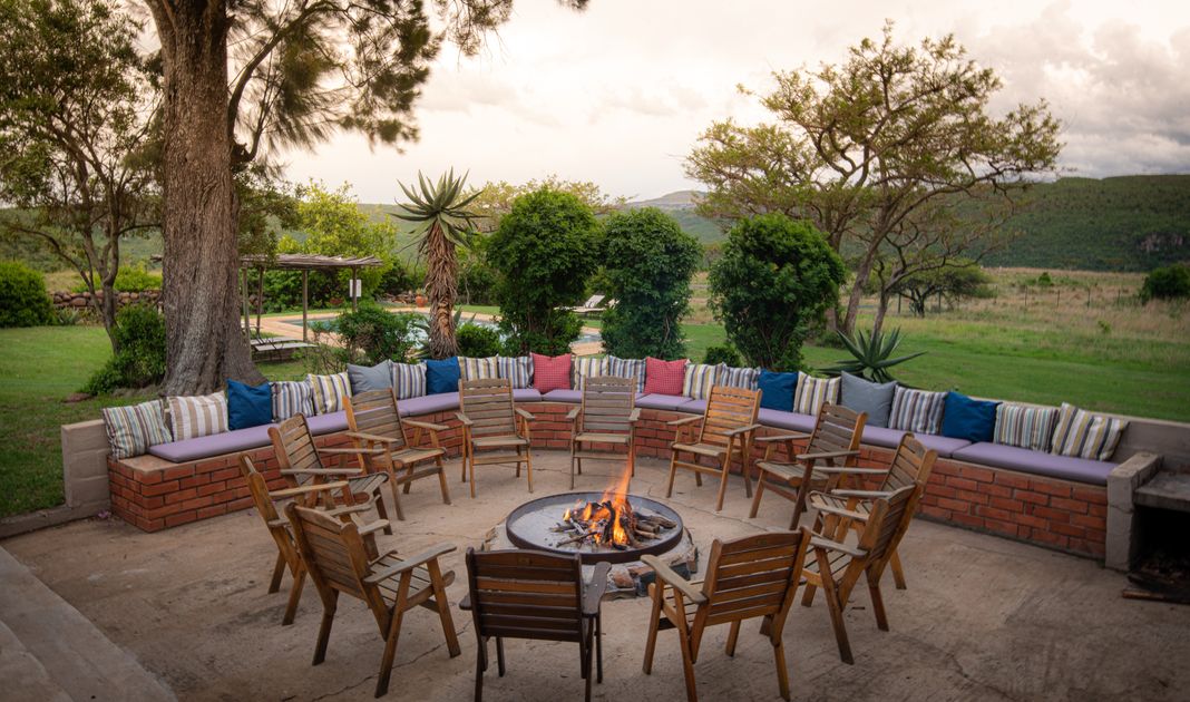 A brick fire-circle bench with cushions around a lit fire pit on a patio, with a garden, pool, and green reserve hills beyond.