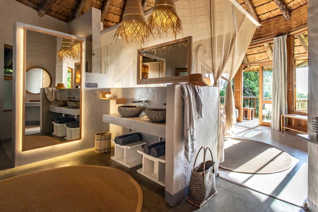 An en-suite with twin stone basins under a lit full-length mirror, opening onto a deck.
