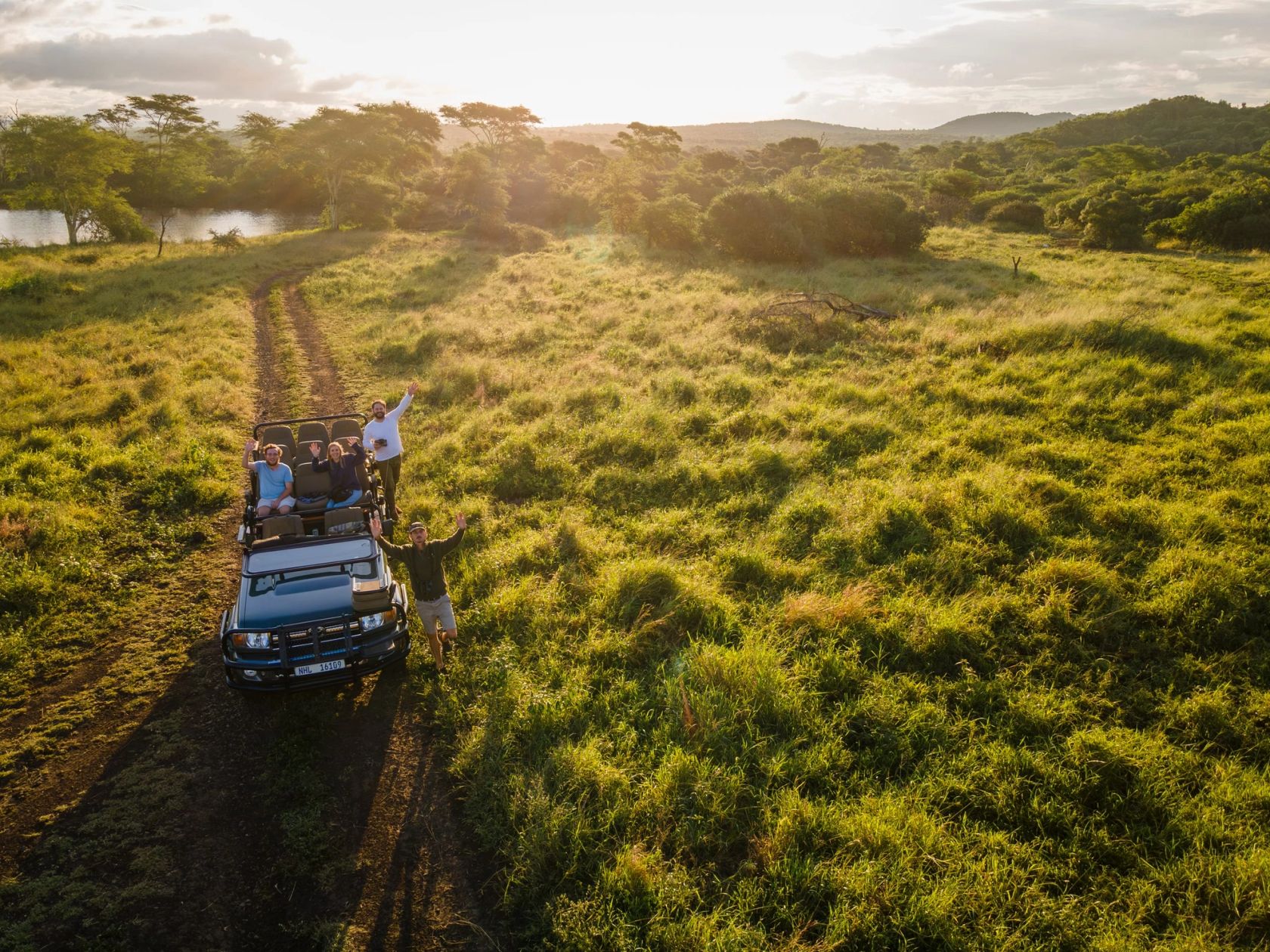 An open safari vehicle stops on a grassy track at sunset as several people stand and wave beside it.