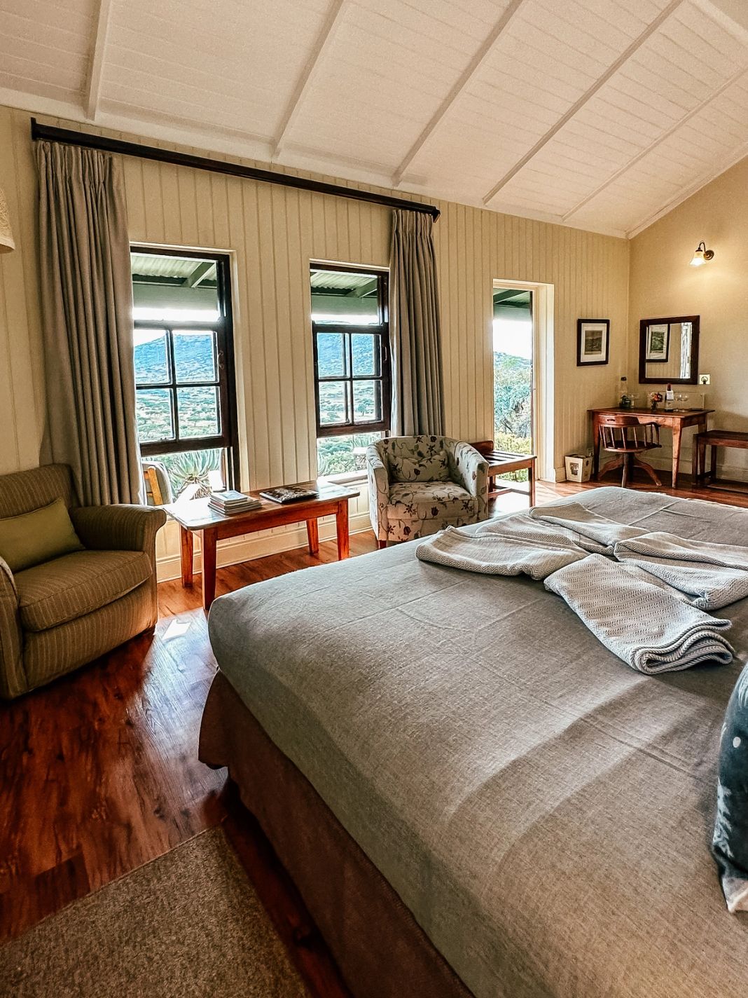 A cottage bedroom with a wide grey-linen bed, two armchairs and tall windows looking across veld to a distant hill.