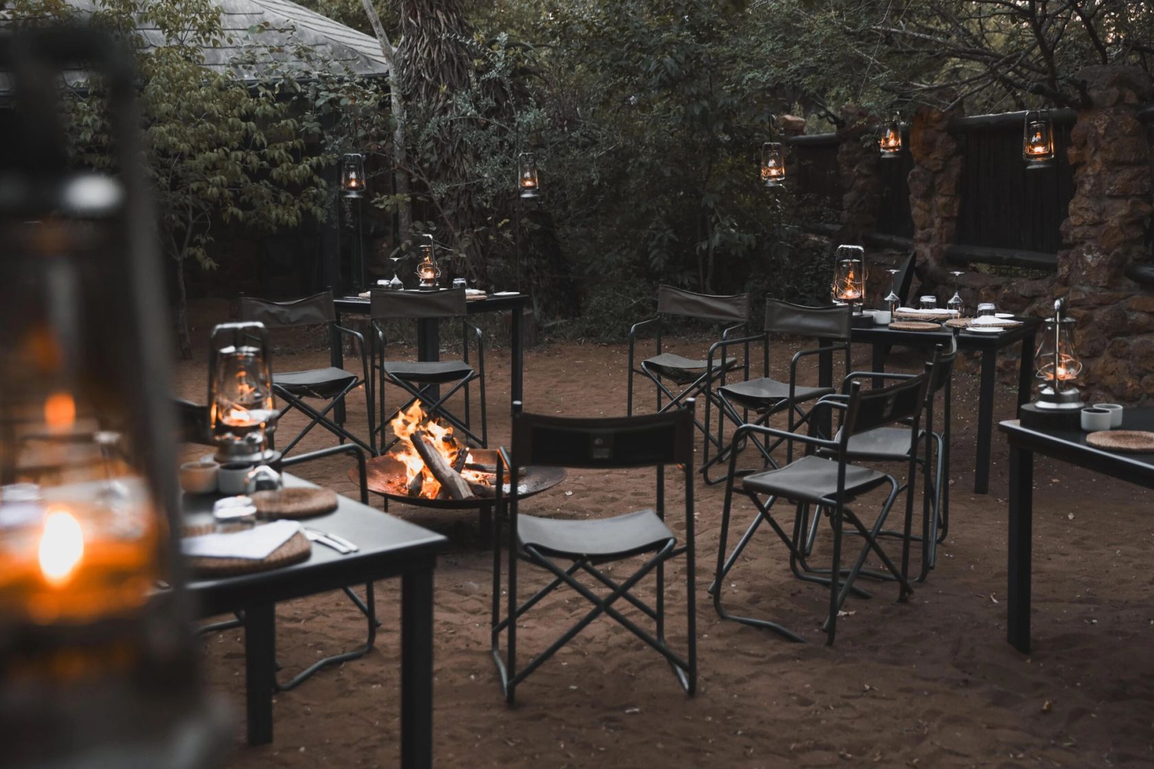 Open-air boma dining tables set with lanterns around a lit fire pit at dusk under trees.