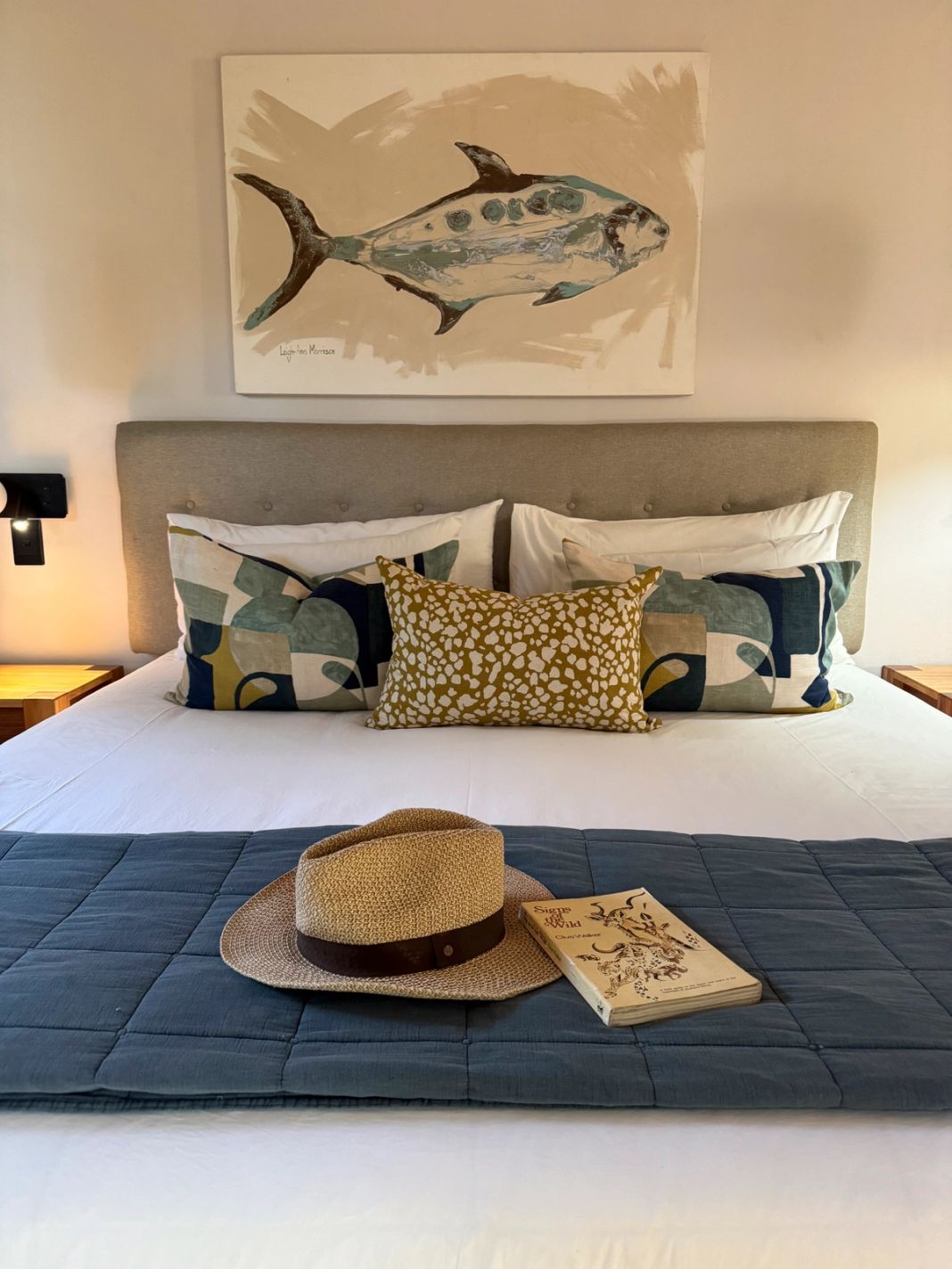 A luxury suite bed beneath a fish painting, with a sun hat and a book laid on the blue quilt.