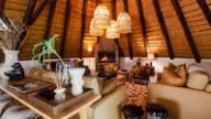 A console holds a vase and reed diffuser in a thatched lounge with sofas gathered around a stone fireplace.