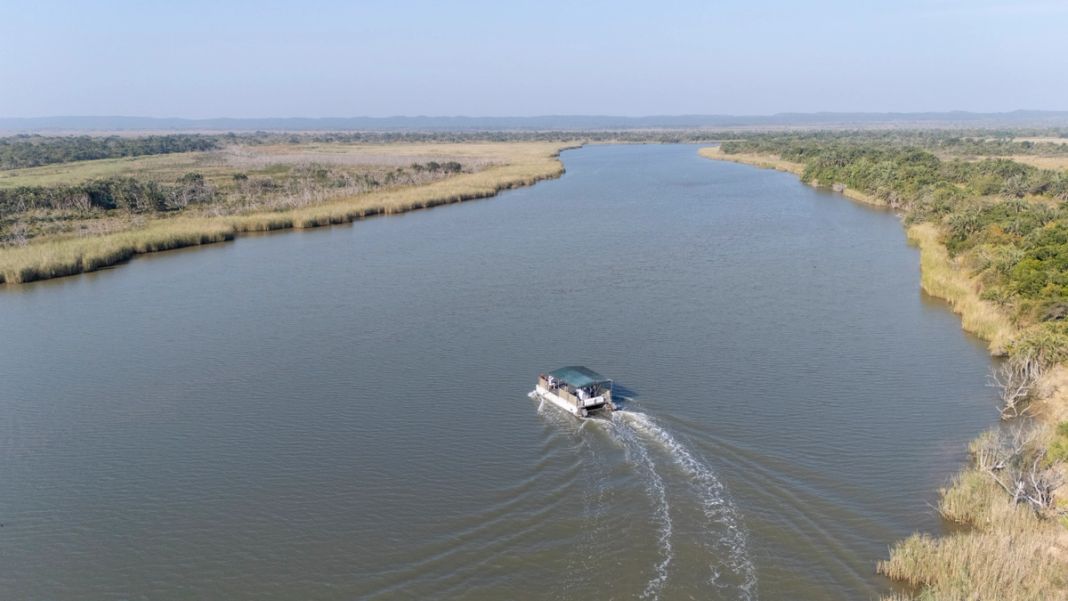 A cruise boat crosses a broad channel of the Lake St Lucia estuary between reed-lined banks.