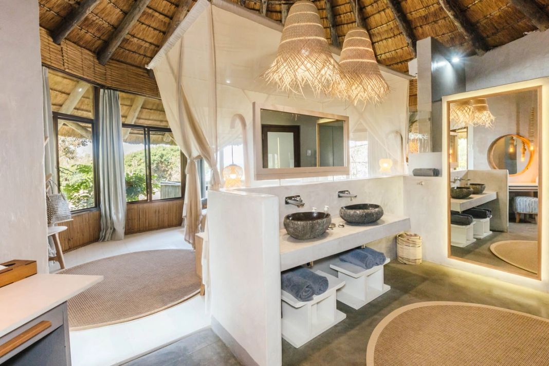 A bathroom with twin stone basins and a window framing green forest bush.
