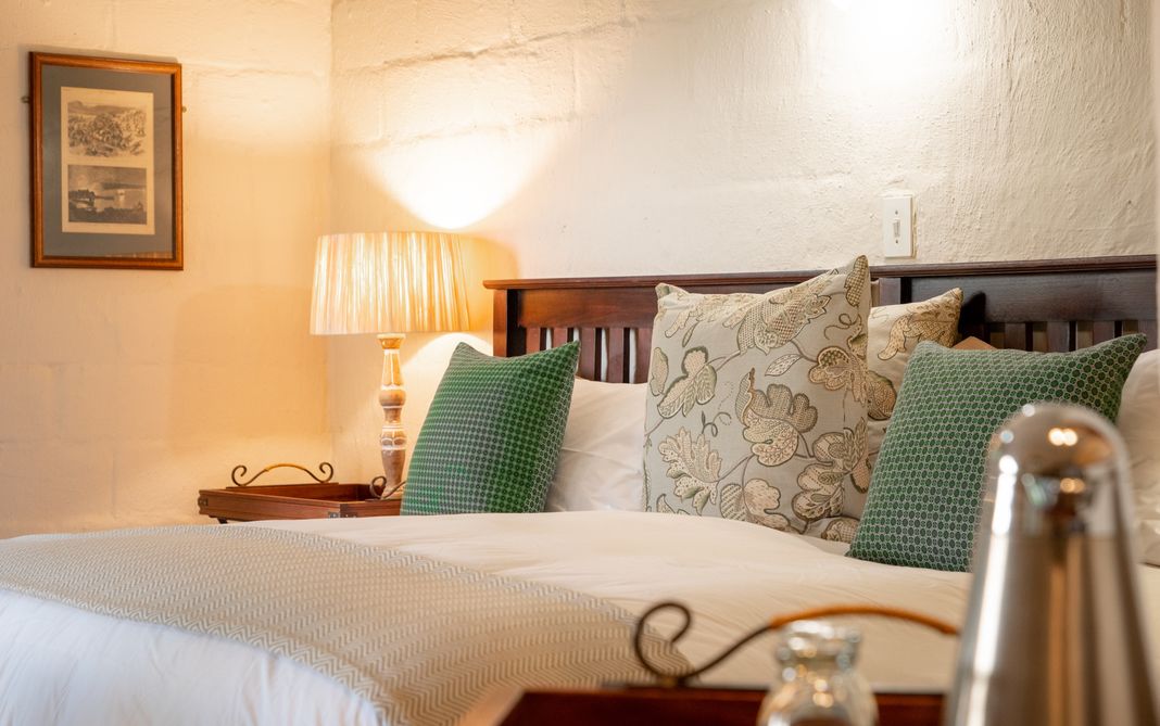 A bed dressed in white linen with green and floral cushions against a whitewashed stone wall, beside a lamp and a framed engraving.