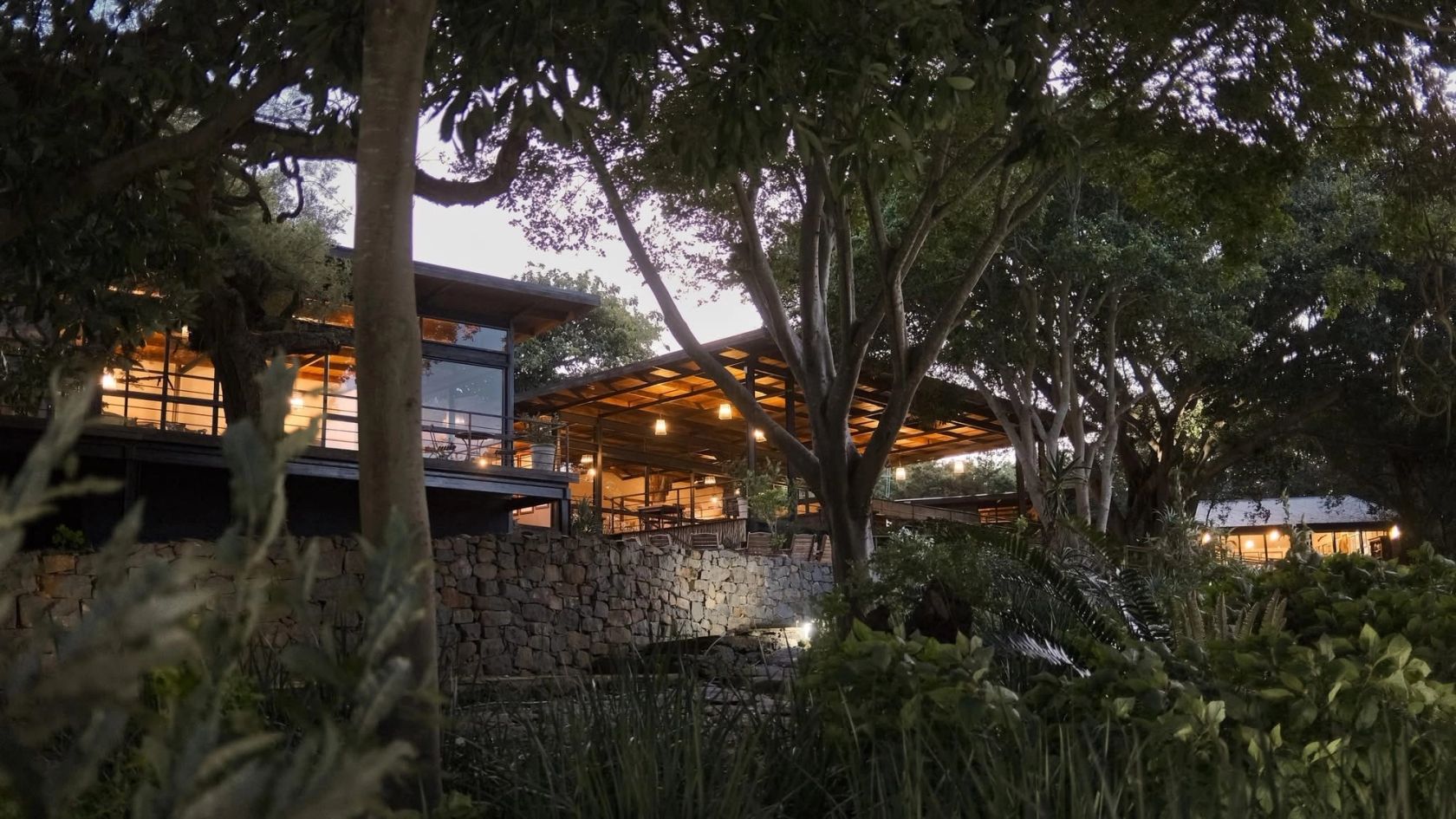 The lodge main building lit from within at dusk, seen through garden trees above a dry-stone wall.