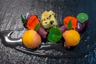 A plated appetizer of cured meat, melon, and a sesame-crusted fritter on dark slate.