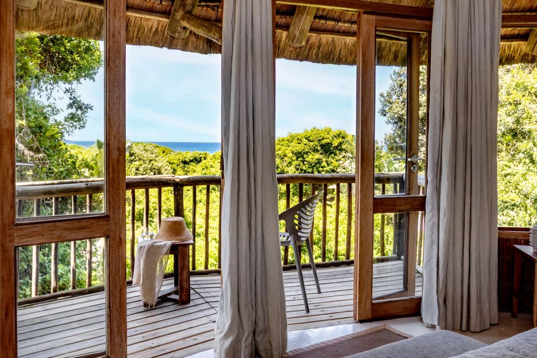The view from the bed through open glass doors to the private deck, a thin band of sea over the forest.