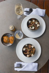 Two plates of beef and feta salad with bread rolls, white wine and water viewed from above on a stone table.