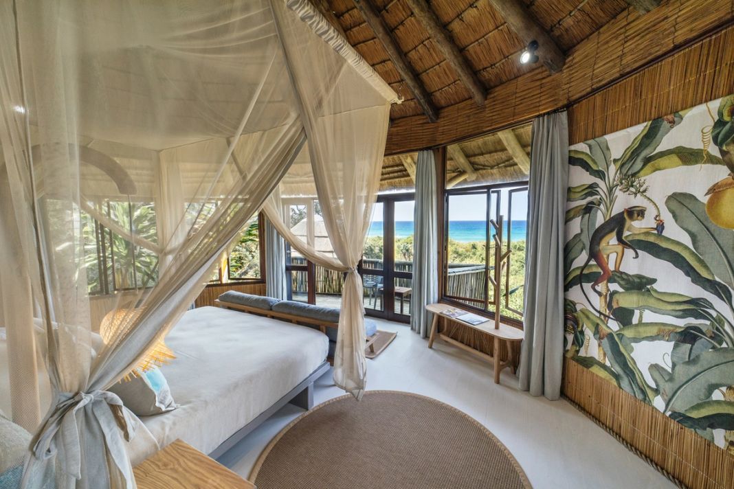 A four-poster bed under a mosquito net, corner windows framing the Indian Ocean beyond the dune forest.