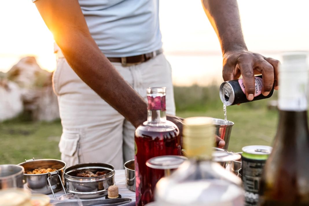 A drink is poured at a table of bottles and snacks beside the water at golden hour.