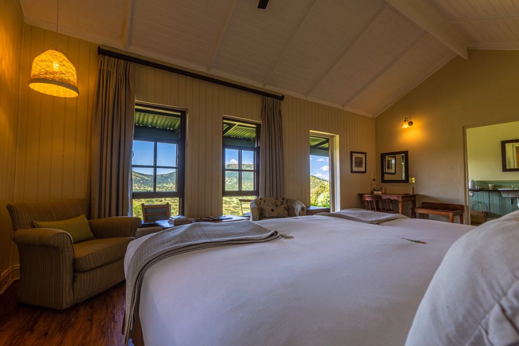 A cottage bed faces three windows framing green rolling hills, with striped armchairs, a woven pendant light and an en-suite visible at right.